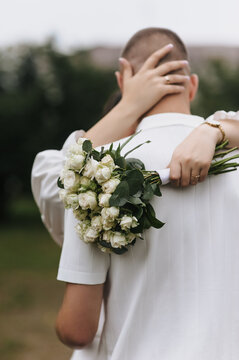 Wedding photo of a bride holding a bouquet of roses and hugging her groom from behind outdoors.