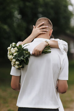 Photo of a bride holding a bouquet of roses and hugging her groom from behind outdoors.