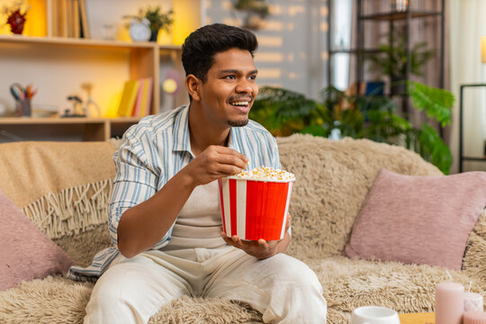 Indian man on home sofa eats popcorn and watches exciting movie with wide eyes and joyful surprise. Arabian guy on home sofa enjoys fun screen time and celebrates thrilling scene in cozy living room.