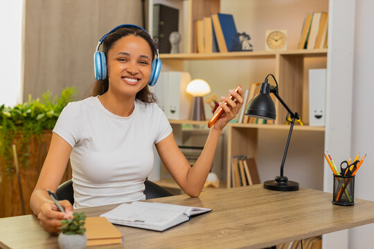 African American young woman on home table takes language test on smartphone and sees right answer. Black girl makes notes in notebook while studying words with headphones in an online lesson at home.