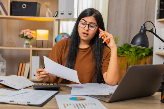Indian woman on home table finds a billing error and calls support to discuss a wrong charge on receipt. Arabian girl checks account details during phone call and asks for payment correction at home.