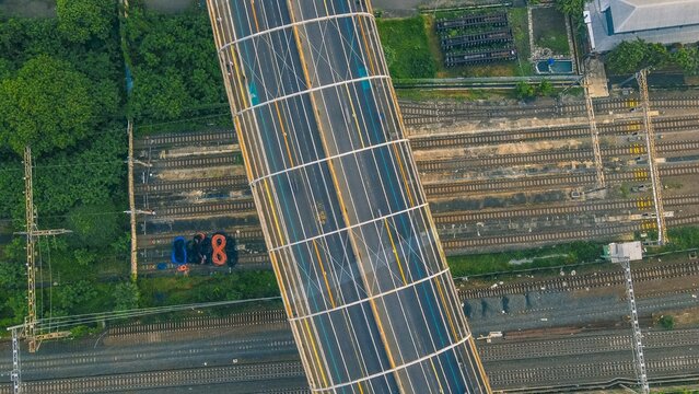 Aerial view of an overpass crossing over multiple railway tracks with overhead power lines and lush green vegetation in Bekasi Barat, West Java, Indonesia.