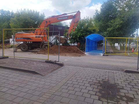 Karaganda, Kazakhstan, 10.09.2026: Excavator behind yellow construction barrier, next to pile of soil and blue covered pedestrian walkway. Roadworks on Voinov-Internatsionalistov Street. Dogsan logo.