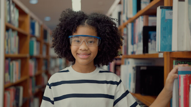 A smiling girl with curly hair and safety glasses stands in a library aisle, wearing a striped t-shirt and reaching for a book on the shelf