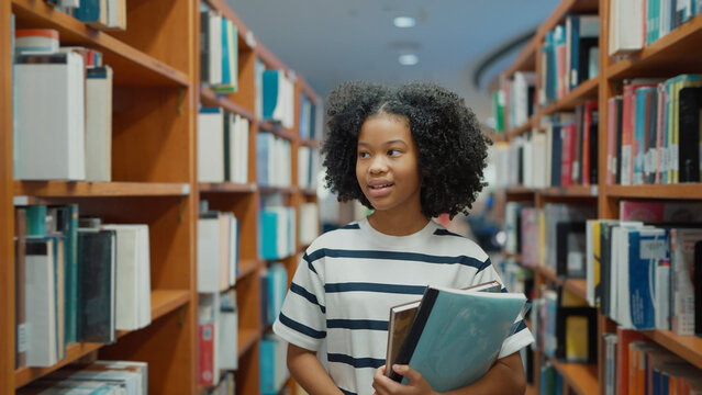 A young girl with curly hair and a striped t-shirt walks down a library aisle, carrying books and folders while looking at the shelves