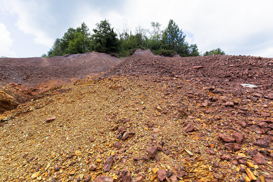 A vast slope of red and orange mineral gravel rises toward green trees under a cloudy sky at Le Roste di Montieri. These barren badlands reveal the deep geological impact of ancient Tuscan mining.