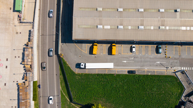 Aerial view of the Esselunga Spa logistics facility with a large warehouse building, parking area with trucks and cars, and adjacent road in Sesto Ulteriano, Lombardia, Italy.