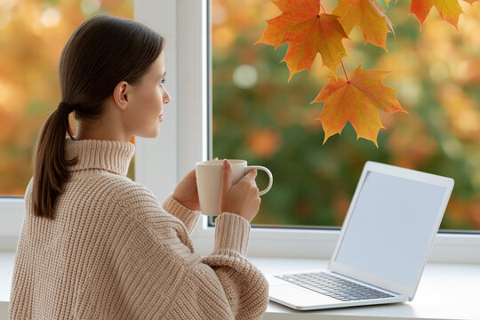 Woman with brown hair in a cozy sweater holding a cup of coffee while sitting by a window with autumn leaves and a laptop on the table