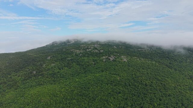 Thick fog rolls slowly over forested summit of Mount Monadnock, exposed granite rocks dot dense green canopy, clear blue sky opens above cloud line in New Hampshire. Aerial drone wide shot