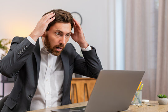 Middle-aged businessman at home office watches laptop freeze eyes widen and presses keys to save work before reboot. Freelancer guy at table restarts system worries about data loss and backup plan ok.