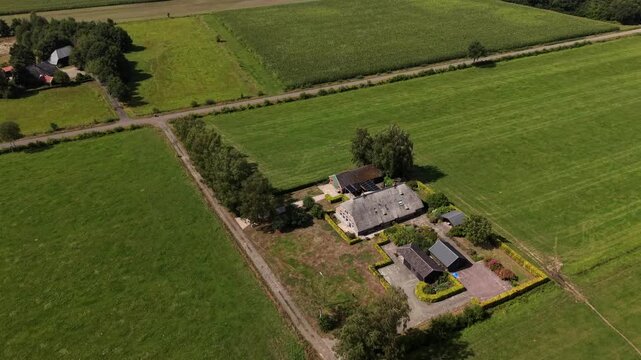 Farmhouse and outbuildings sit isolated among patchwork green fields in Netherlands, straight dirt lane divides crop rows, hedgerows trace rural property boundaries. Aerial drone top down flyover