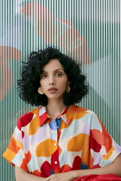 Female model with curly black hair wearing a colorful patterned shirt poses against a textured background with abstract shapes and lines in a modern setting