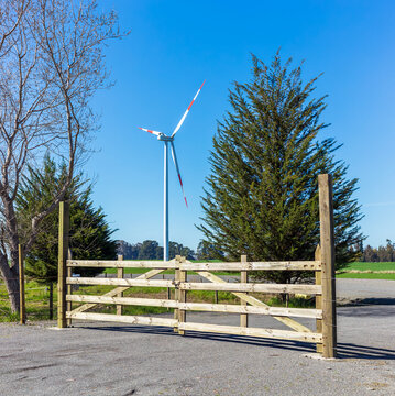 A rustic wooden gate opens toward green fields and a tall wind turbine in the Biob&iacute;o Region of Chile, set beneath a clear blue sky
