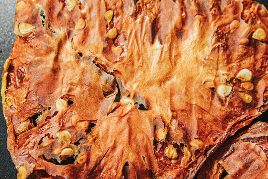 Extreme close-up of a dried tomato slice showing translucent fibrous flesh, wrinkled red-brown skin, and clusters of golden seeds against a dark background.