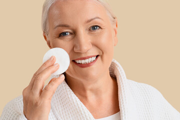 Healthy aging and skincare routine, senior woman using cotton pad for facial cleansing in bathrobe for wellness and beauty self care on beige background © Pixel-Shot