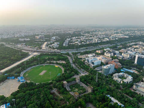 Aerial view of a cricket ground surrounded by lush green trees and modern office buildings in the urban landscape of Gurgaon, Haryana, India.