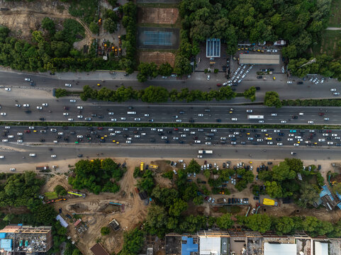 Aerial view of a busy multi-lane highway with heavy traffic flow, surrounding green trees, and urban development in Gurgaon, Haryana, India.