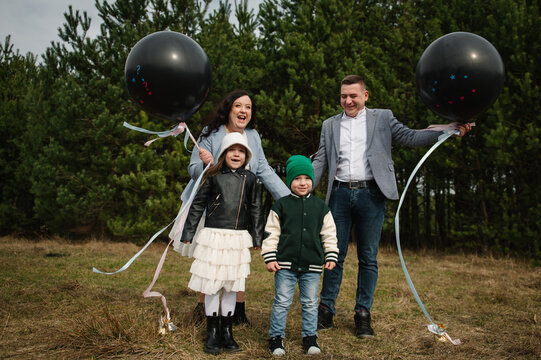 Happy family of four with two children holding black gender reveal balloons in a pine forest during spring outdoor photoshoot.