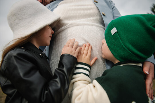 Cute children gently touching and kissing the belly of their pregnant mother outdoors during a family twin pregnancy photoshoot.