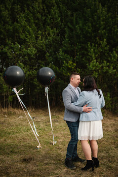 Loving pregnant woman and her husband embracing tenderly outdoors in a forest during a twin gender reveal celebration with two black balloons.