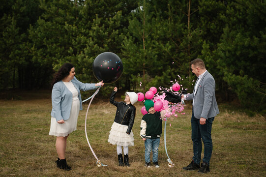 Happy family popping a black gender reveal balloon with pink confetti bursting out during an outdoor celebration in a forest for twins.