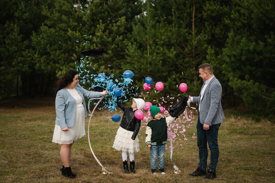 Happy family popping both gender reveal balloons with blue and pink confetti bursting out during a twin celebration in a forest.