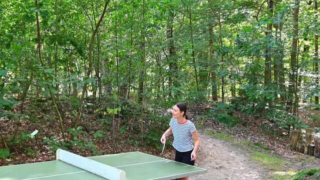 A candid moment of a young woman experiencing a series of humorous, unsuccessful attempts at a friendly, amateur table tennis (ping-pong) match outdoors in a scenic forest setting.
