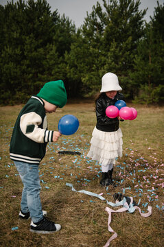 Little boy and girl playing with small blue and pink balloons among scattered confetti in a pine forest.