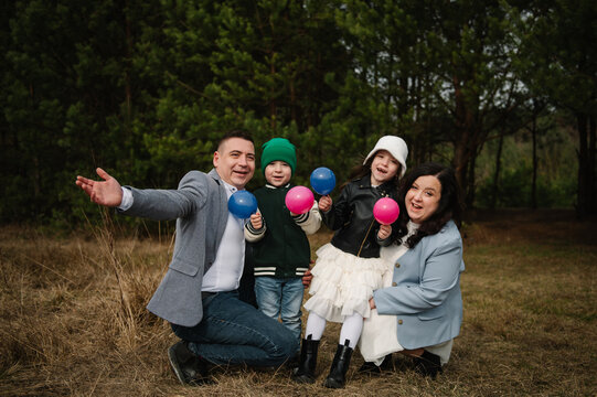 Happy family with children holding small blue and pink balloons posing outdoors in a forest during a gender reveal celebration.