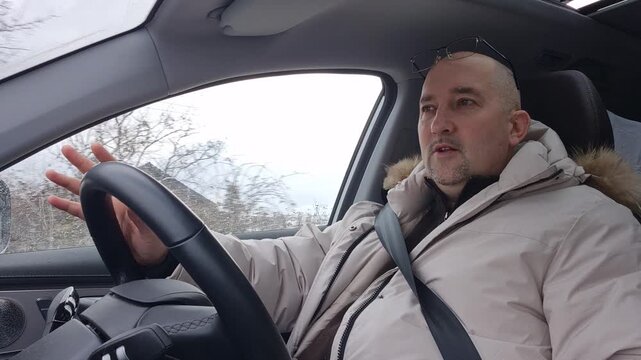 Middle aged man driving car through snowy town, gesturing while speaking to passengers. Fixed interior camera shot.