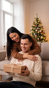 Couple celebrating Christmas together, woman embraces man as he opens a gift in a cozy living room with a decorated tree