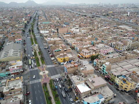Aerial view of Avenida Peru featuring a wide multi-lane road with traffic, surrounded by dense urban residential buildings and rooftops in San Martin de Porres, Lima, Peru.