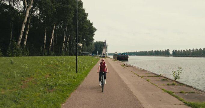 Front view drone tracking a woman leisurely cycling along a bike path beside a forest and river, enjoying nature while using her bicycle for transportation.