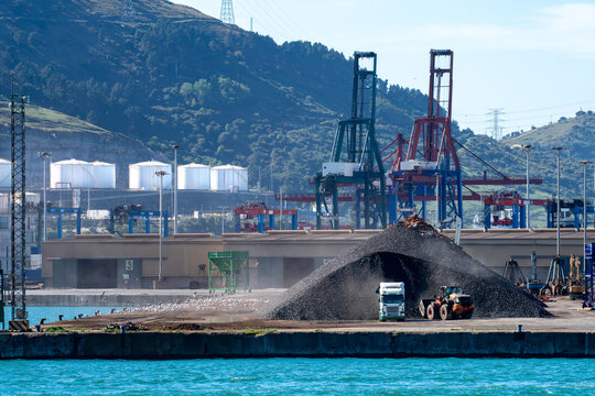 View of Getxo port with a large pile of coal, a truck, and a front loader operating near industrial cranes and storage tanks in Biscay, Basque Country, Spain.