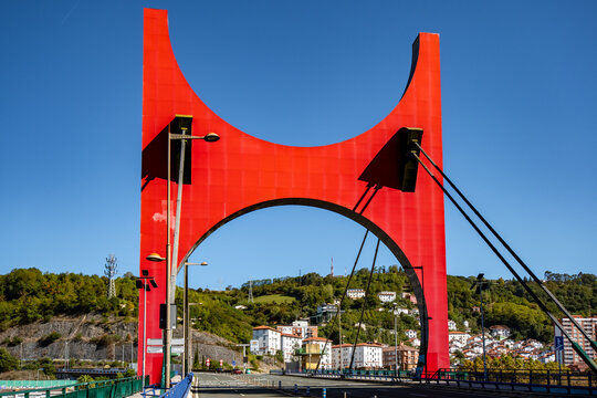 View of the iconic bright red arch of the La Salve Bridge spanning the Nervion River, with the surrounding urban landscape and green hills of Bilbao, Basque Country, Spain.