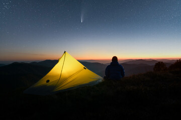 Yellow tent and hiker resting on a mountain ridge beneath a star-filled night sky. Faint aurora and distant mountains create a peaceful wilderness camping scene © Ivan Kmit