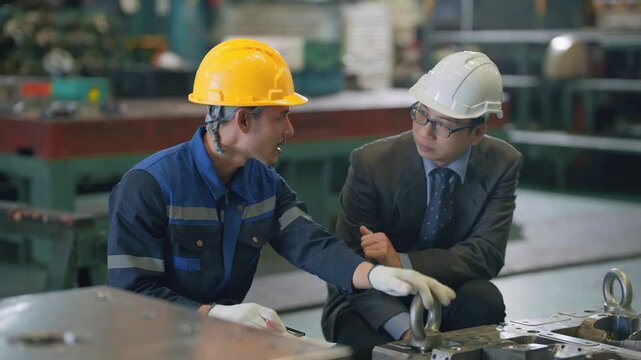 Two men in hard hats inspect machinery together. One wears blue work clothes, the other suits. They gesture and point, focused on the task