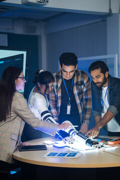 A group of diverse professionals working together in a modern workspace, gathered around a table to test and inspect a robotic arm prototype during a late shift project development session.
