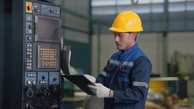 Worker wears hard hat and safety gloves. He stands near industrial machine controls. He checks documents while looking at display panels
