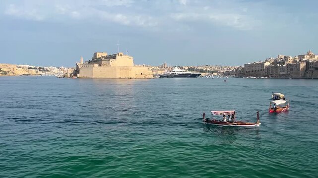 Scenic view of Grand Harbour in Malta with historic Fort St. Angelo and Birgu waterfront in the background. Traditional Maltese dgħajsa boats cross the turquoise water on a sunny summer day