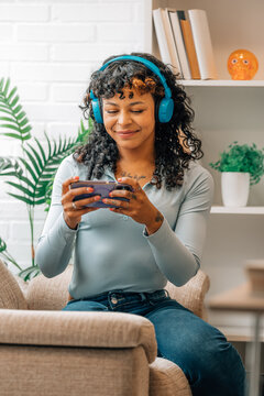 Smiling woman wearing wireless headphones and using smartphone at home, enjoying music or video content while relaxing on a couch in a bright
