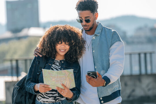 Young African American couple exploring the city together, smiling woman reading a paper map while man checks smartphone, urban travel and tourism lifestyle concept outdoors