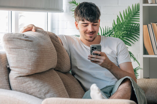 Smiling young man relaxing on sofa at home, using smartphone, casual lifestyle indoors