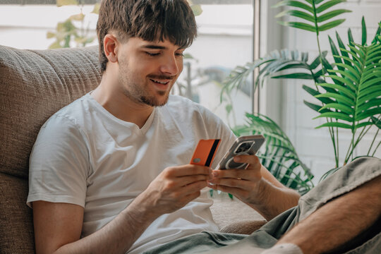 Smiling young man sitting on a sofa at home, holding a credit card and smartphone, shopping online or making a mobile payment