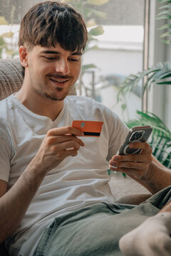 Smiling young man sitting on a sofa at home, holding a credit card and smartphone, shopping online or making a mobile payment