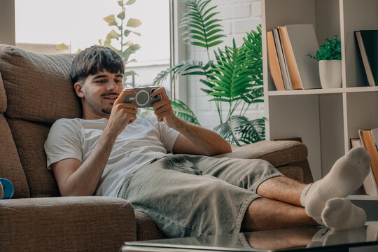 Young Caucasian man relaxing in an armchair at home, smiling while playing a game on his smartphone, enjoying leisure time in a cozy living room