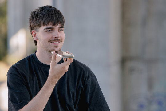 Young Caucasian man smiling while recording a voice message on his smartphone, holding the phone close to his mouth outdoors
