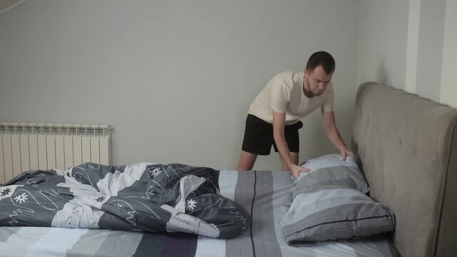 Young man smoothing duvet and arranging pillow while making bed in minimalist bedroom with tufted gray headboard and soft daylight neat bedding folds and calm housekeeping routine