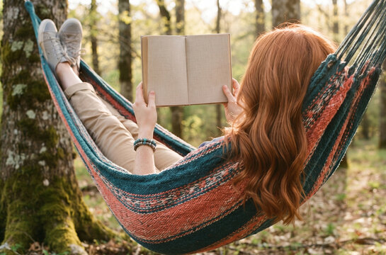 Red-haired woman reading a book in a colorful hammock in a forest