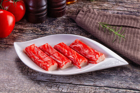 Four red and white imitation crab sticks on white ceramic plate with fresh tomatoes, rosemary sprig, and wooden background